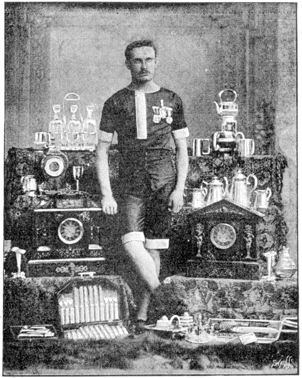 Ein Schwarz-Weiß-Foto eines Mannes in einem Fussballtrikot vor einem Tisch voller Trophäen und Pokale, mit der Aufschrift "1909-1910 WM-Sieger" darunter.