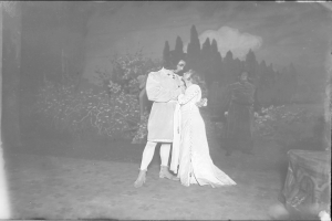 Ein Schwarz-Weiß-Foto eines Mannes und einer Frau, die sich auf einer Bühne umarmen und küssen, mit Menschen, Pflanzen und Bäumen im Hintergrund.