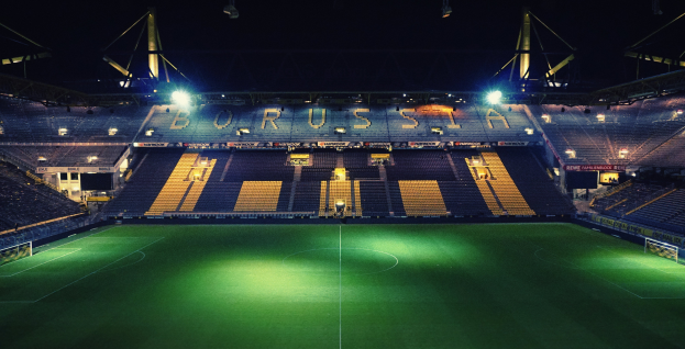 Ein Fubballfeld nachts, erleuchtet von hellen Stadionlichtern, mit ein paar Menschen auf den R├Ąngen und saftig gr├╝nem Rasen.