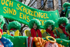 Eine Gruppe von Clowns in bunten Kostümen marschiert, eine Fahne mit Text haltend, mit Bäumen und einem Gebäude im Hintergrund.