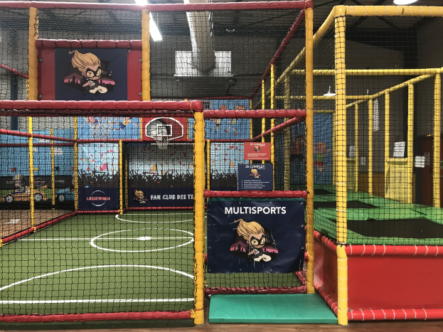 Großes Indoor-Trampolinpark mit einem Basketballfeld in der Mitte, umgeben von einem Netz, mit einem Basketballkorb, Wandbannern und Deckenleuchten.