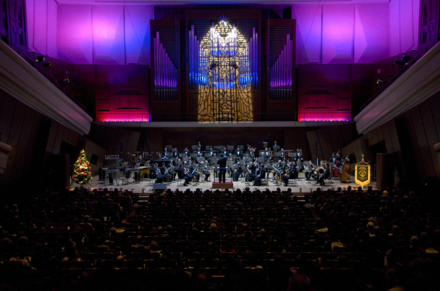 Ein großer Saal voller Menschen bei einem Konzert, mit Musikern auf der Bühne und einem geschmückten Weihnachtsbaum im Hintergrund.
