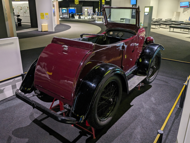 Alter roter Wagen in einem Museum mit Absperrpollern, Bändern, Informationsschildern und umgebenen Ausstellungsstücken.