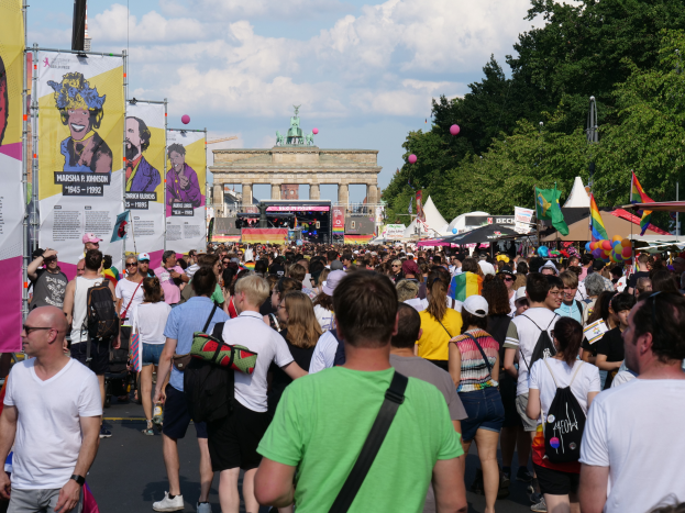 Eine große Menschenmenge marschiert vor dem Brandenburger Tor in Berlin, Deutschland, mit vielen, die Mützen und Taschen tragen, Schilder auf der linken Seite und im Hintergrund Bäume, Zelte, Luftballons, ein Tor und ein bewölkter Himmel.