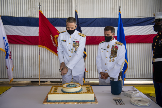 Zwei Männer in weißen Marineuniformen schneiden einen Kuchen an einem Tisch mit Besteck und einem Eimer, mit Fahnen und einem Metallblech im Hintergrund.