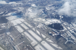 Luftaufnahme eines schneebedeckten Flughafens mit Gebäuden, Straßen, Brücken, Wasser, Bäumen und Wolken.