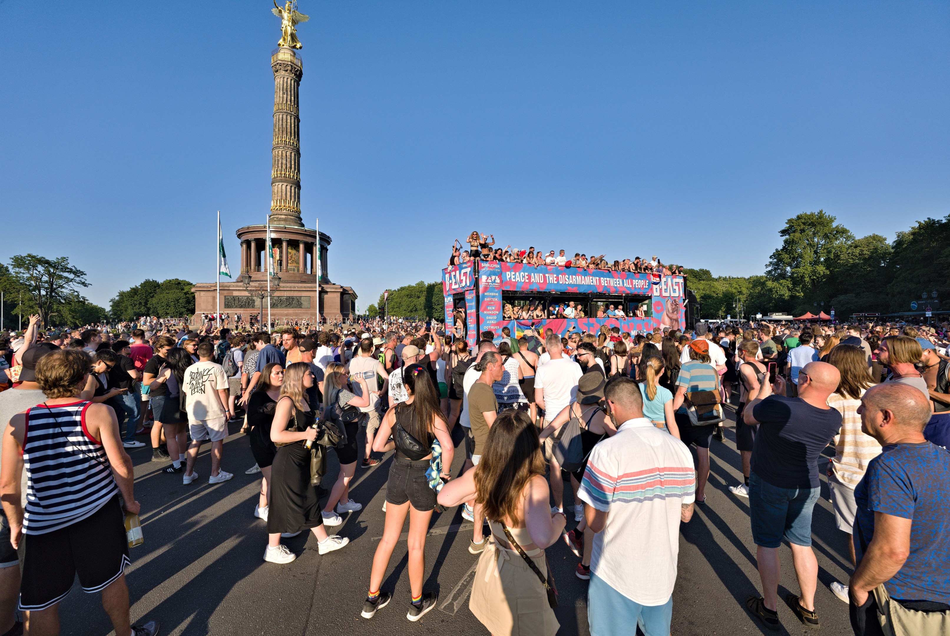 Große Menschenmenge vor einem Denkmal in Berlin, Deutschland, mit Fahnen, einem Gebäude mit Säulen und einer Statue, Bäumen und einem klaren blauen Himmel.