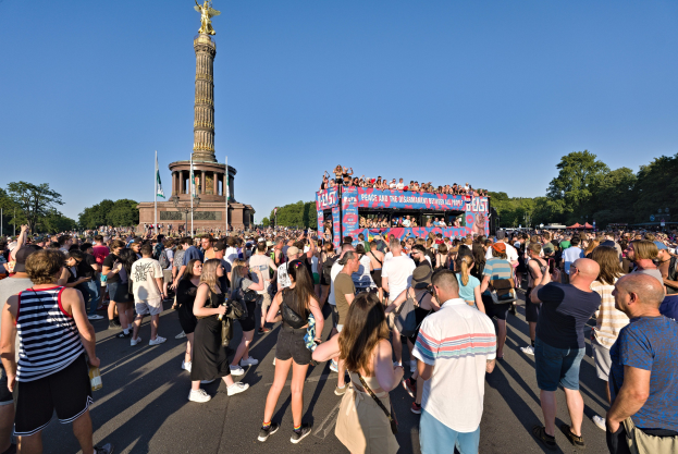 Große Menschenmenge vor einem Denkmal in Berlin, Deutschland, mit Fahnen, einem Gebäude mit Säulen und einer Statue, Bäumen und einem klaren blauen Himmel.