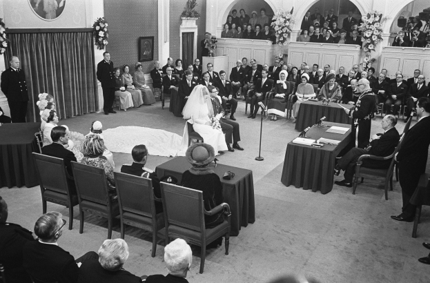 Schwarzes und weißes Foto einer Hochzeitszeremonie in einem großen Raum mit sitzenden und stehenden Gästen, Tischen mit Mikrofonen und Papieren, einer hinteren Wand mit Rahmenfotos, Blumenbouquets, einem Vorhang und einem Podium mit Mikrofonständer.