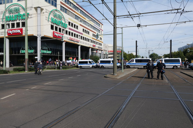 Eine Gruppe von Polizisten steht auf der Straße neben einer Tram, mit Fahrzeugen, Strommasten, Gebäuden, Bäumen und einem bewölkten Himmel im Hintergrund.