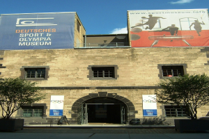 Außenansicht des Deutschen Sport & Olympia Museums in Berlin, das ein Gebäude mit Fenstern und einer Tür zeigt, umgeben von Bäumen und Bannern unter einem bewölkten Himmel.