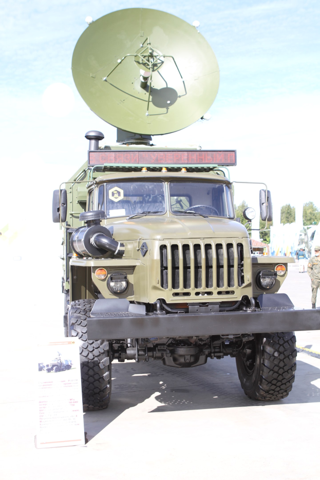 Militärlastwagen mit montierter Satellitenschüssel, auf einem Feld mit Menschen, Bäumen und einem klaren blauen Himmel geparkt.