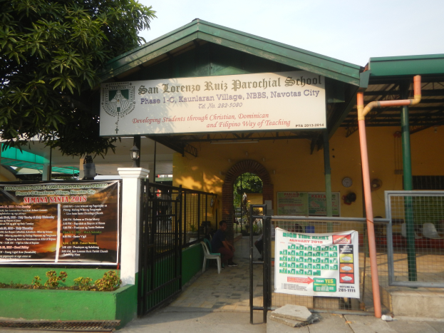 Der Eingang der San Lorenzo Ruiz Pfarrschule hat eine Tafel mit Text, einen Metallzaun, Menschen, die unter einem Dach sitzen, Pfosten, ein Schild, Pflanzen, einen Baum und einen bewölkten Himmel.