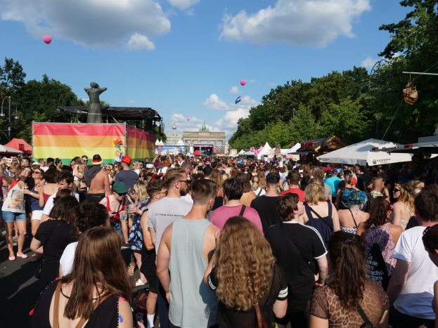 Eine große Menschenmenge marschiert auf einer Straße mit Zelten, Bäumen, Pfählen, Lichtern und einer Statue im Hintergrund, mit Gebäuden und einem Himmel mit Wolken und Ballons, während des Christopher Street Day in Berlin.