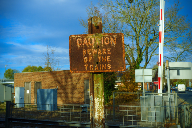 Vorsichtsschild an einer Bahnschiene umgeben von einem Metallzaun, Bäumen, Strommasten, einem Gebäude, Containern, Fahrzeugen auf einer Straße und einem bewölkten Himmel.