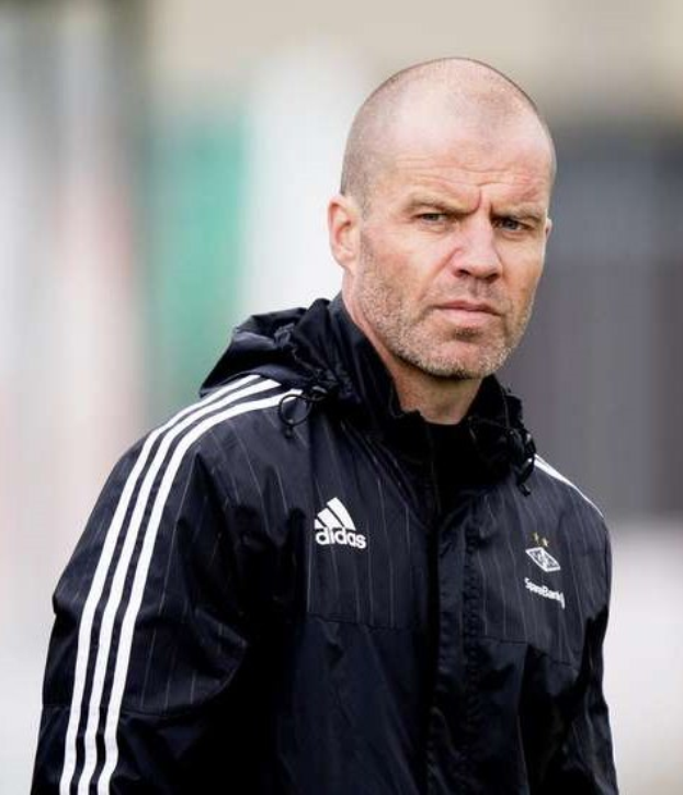 Ein kahlköpfiger Mann in einer schwarzen Jacke, der wie ein Fußballtrainer aussieht, steht auf einem Feld mit gefalteten Händen und einem ernsten Gesichtsausdruck, trägt eine blaue Jacke mit weißen Streifen und einem weißen Kragen.