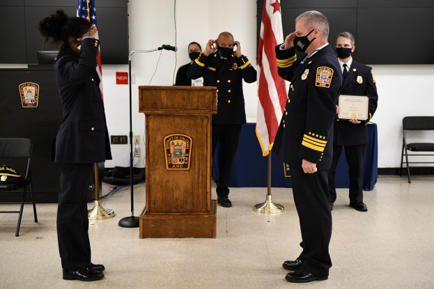 Uniformierte Personen in Masken stehen vor einem Podium mit Mikrofon während einer Vereidigung eines neuen Feuerchefs, haltend ein Zertifikat, umgeben von Flaggen, Stühlen, Tischen und einer Wand.