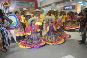 Eine Gruppe von Menschen in bunten Kleidern und Hüten, die in einem Einkaufszentrum tanzen, während andere Musikinstrumente halten und ein Banner im Hintergrund zu sehen ist.