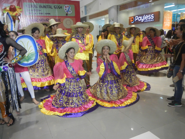 Eine Gruppe von Menschen in bunten Kleidern und Hüten, die in einem Einkaufszentrum tanzen, während andere Musikinstrumente halten und ein Banner im Hintergrund zu sehen ist.