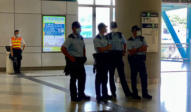 Eine Gruppe von Polizisten in Masken steht vor einem Gebäude mit Glastüren, Schildern mit Text, einer Säule und einem Geländer, wobei durch die Glastüren Bäume, Fahrzeuge und andere Gegenstände zu sehen sind, was darauf hinweist, dass sie in Hong Kong verhaftet wurden.