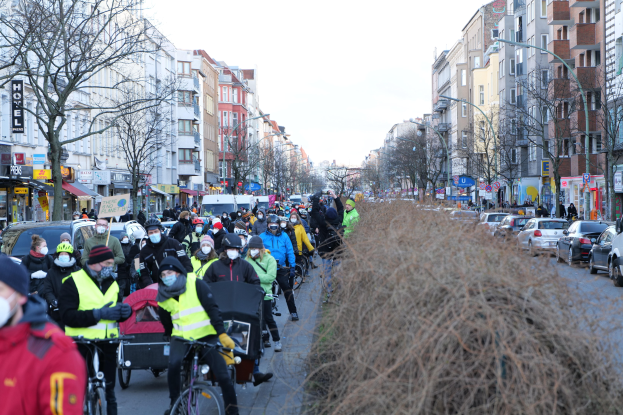 Eine große Gruppe von Menschen in Masken und Sicherheitswesten auf Fahrrädern eine von Bäumen gesäumte Straße entlangfahren, begleitet von Fahrzeugen und trockenem Gras, unter einem klaren blauen Himmel.