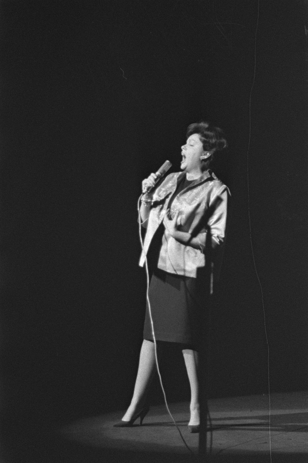 Eine Frau in einem schwarzen und weißen Outfit aus den 1950er-Jahren singt fröhlich auf einer von einem Scheinwerfer beleuchteten Bühne und hält ein Mikrofon in der Hand.