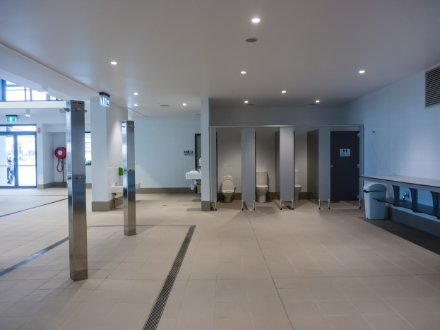 Großes öffentliches WC mit mehreren Toiletten, tragenden Säulen, einem Mülleimer, einer Tür, einem Schild, einem angebrachten Feuerlöscher und Deckenleuchten.