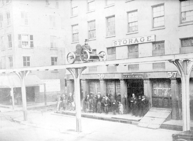 Altes Schwarz-Weiß-Foto eines Feuerwehrautos, das vor einem Gebäude mit einer Namensplakette geparkt ist, eine Person auf dem Fahrzeug sitzt, eine Gruppe von Menschen in der Nähe steht und ein Hund vorhanden ist.