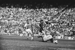 Schwarzes Foto von Fußballspielern im Einsatz auf einem Feld mit Zuschauern im Hintergrund und einer Bundesliga 1978-79 Ajax 1-1 Hannover 96-Fahne.