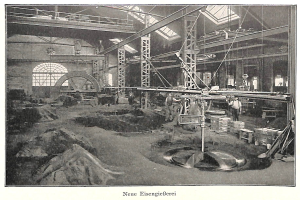 Schwarzes und weißes Foto eines Fabrikinneren mit Arbeitern, die Maschinen bedienen, umgeben von Metallstangen und -pfosten, beschriftet mit "Neue Eisengießerei, die erste Fabrik Deutschlands" unten.