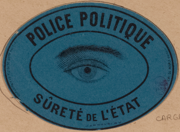Eine braune Kartonschachtel mit einem blauen Aufkleber, auf dem "Polizei Politique Sûreté de l'État" und ein Auge in der Mitte steht.