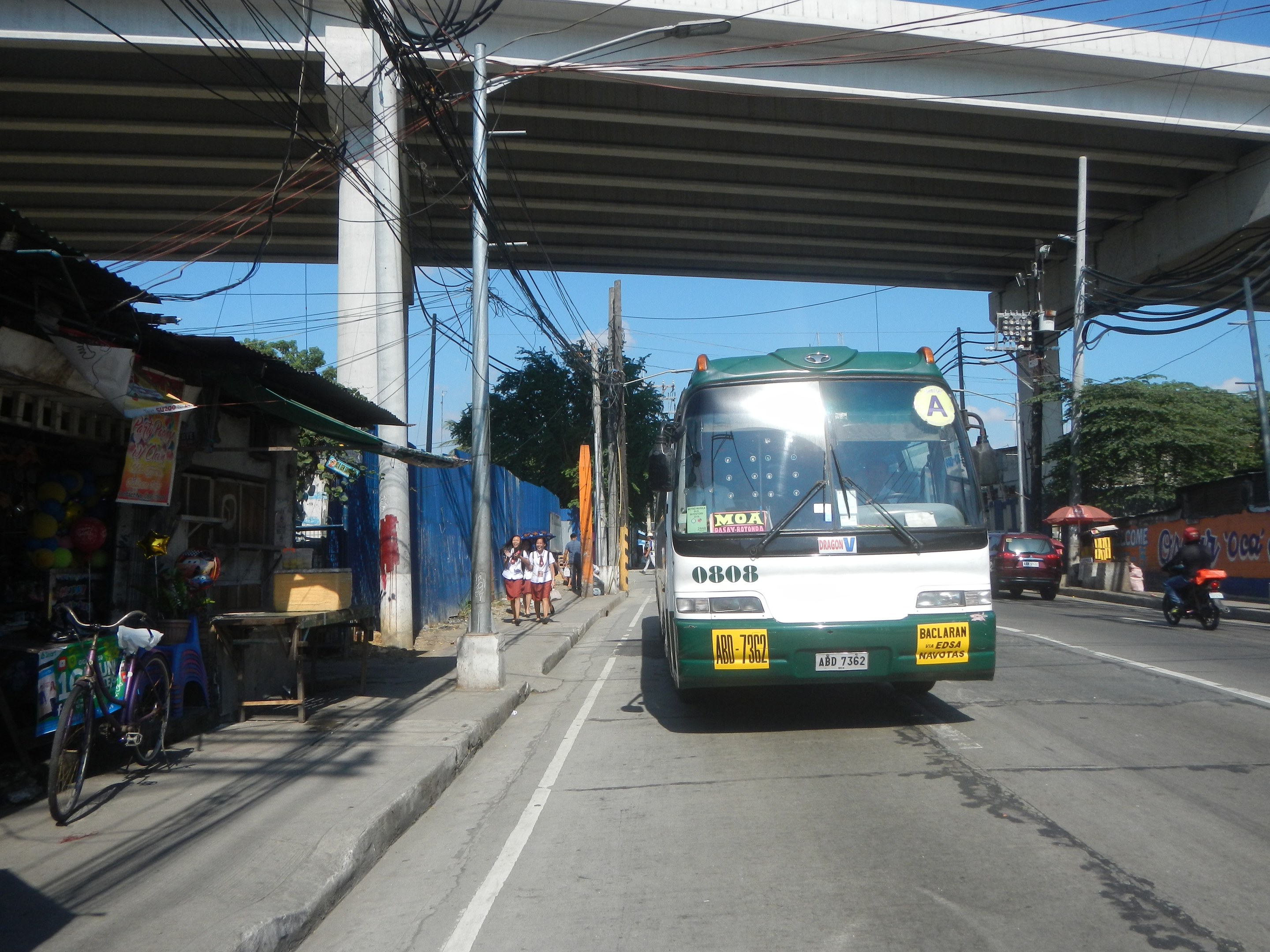 Ein grüner und weißer Bus fährt eine Straße mit Geschäften und Ständen entlang, unterquert eine Brücke mit Strommasten und -leitungen und fährt an Fußgängern und Fahrrädern vorbei, während sich im Hintergrund Bäume und ein klarer Himmel befinden.