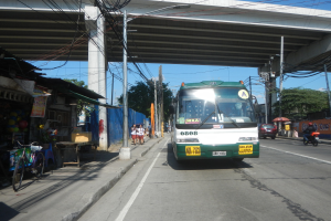 Ein grüner und weißer Bus fährt eine Straße mit Geschäften und Ständen entlang, unterquert eine Brücke mit Strommasten und -leitungen und fährt an Fußgängern und Fahrrädern vorbei, während sich im Hintergrund Bäume und ein klarer Himmel befinden.