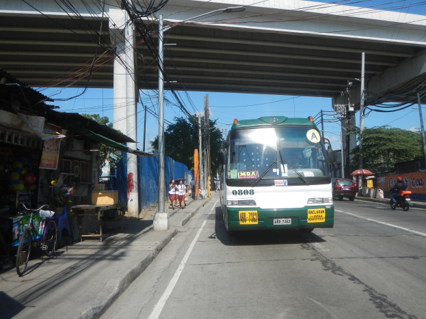 Ein grüner und weißer Bus fährt eine Straße mit Geschäften und Ständen entlang, unterquert eine Brücke mit Strommasten und -leitungen und fährt an Fußgängern und Fahrrädern vorbei, während sich im Hintergrund Bäume und ein klarer Himmel befinden.