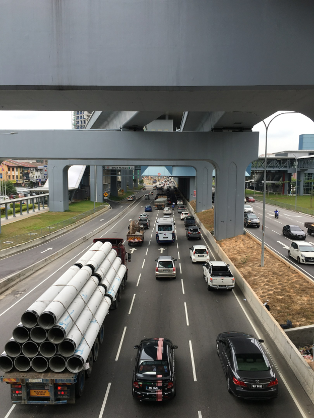Eine befahrene Autobahn mit mehreren Fahrzeugen unter einer Brücke, umgeben von Gras, Bäumen, Gebäuden, Polen und Lichtern, unter einem Himmel.