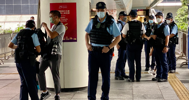 Eine Gruppe uniformierter Polizisten, einige mit Kameras, steht in einer Reihe vor einem Gebäude mit einer Fahne an einer Säule, Geländern, Pflanzen, Bäumen und Deckenleuchten im Hintergrund.