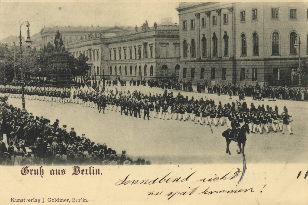 Ein altes Schwarz-Weiß-Foto eines Umzugs in Berlin, Deutschland, mit Menschen auf dem Boden und Pferden, einer Statue auf einem Sockel, Straßenlaternen, Bäumen, Gebäuden mit Fenstern und einem bewölkten Himmel, mit Text am unteren Rand des Bildes.