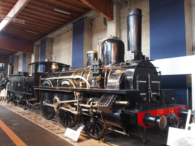 Eine große, alte Dampflokomotive in einem Museums-Setting umgeben von Eisenbahnschienen, Informationsschildern, Ständen und anderen Ausstellungsstücken, mit einer Wand und Deckenbeleuchtung im Hintergrund.