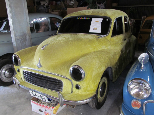 Mehrere Oldtimer sind vor einer Wand mit verschiedenen Accessoires geparkt, wobei auf der linken Seite eine Säule zu sehen ist.