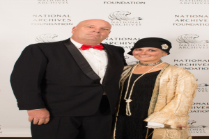 Ein Mann in einem schwarzen Anzug mit einem weißen Hemd und einer schwarzen Fliege steht neben einer Frau in einem eleganten Kleid mit einem gemusterten Schultertuch auf einem roten Teppich, mit einem weißen Banner mit Text im Hintergrund.