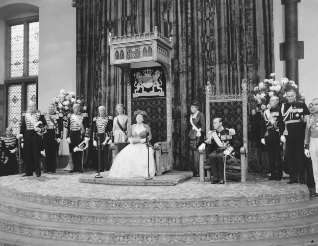 Schwarz-weiß-Fotografie von Queen Elizabeth II., die auf einem Stuhl sitzend in einem weißen Hochzeitskleid und mit einem Blumenstrauß in der Hand zu sehen ist, neben ihr auf der rechten Seite Prinz Philip und mehrere Anwesende auf der linken Seite.