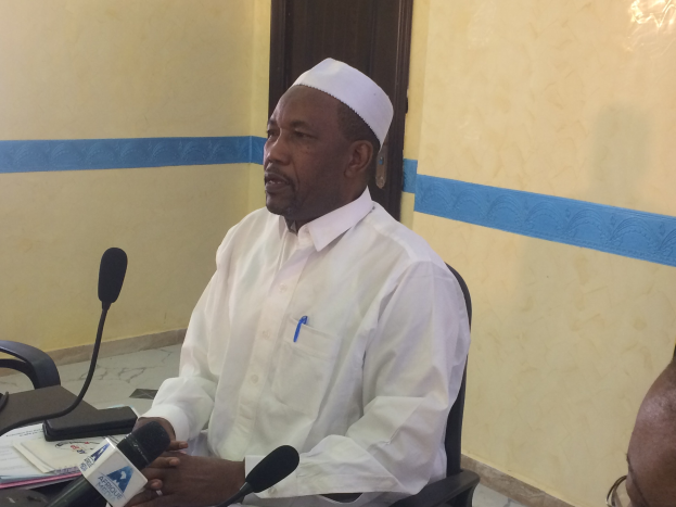 Ein Mann in einem weißen Kleid und einer Mütze sitzt an einem Tisch mit Mikrofonen und spricht, während Papiere und Gegenstände auf dem Tisch zu sehen sind, mit einer anderen Person mit Brille rechts von ihm in einem Raum mit einer Wand und einer Tür im Hintergrund.