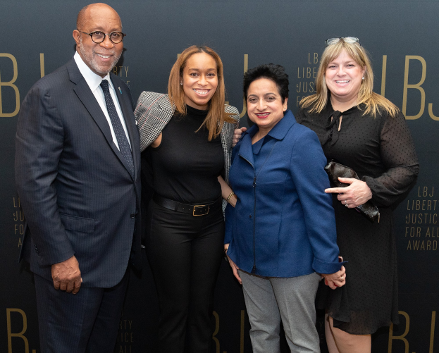 Eine Gruppe von vier Menschen auf einem roten Teppich, die lächeln und posieren, mit einem 'Liberty Justice for All Awards'-Schild im Hintergrund.