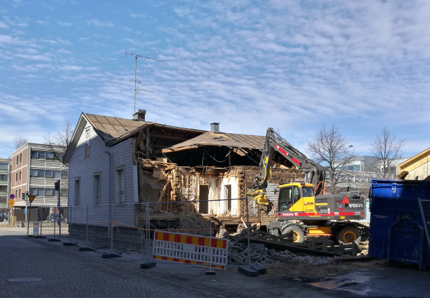 Eine Baustelle mit einem großen Gebäude, das abgerissen wird, mit einem Kran, einem Zaun, einer Absperrung, Steinen, einem Container, Bäumen, Pfählen, einem Schild, einem Straßenpfahl, Fahrzeugen, benachbarten Gebäuden mit Fenstern und einem bewölkten Himmel.
