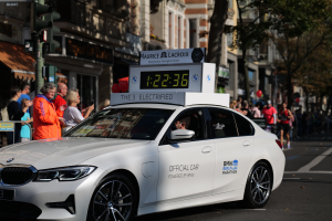 Ein weißer BMW mit einer Uhr auf dem Dach fährt eine Straße entlang, mit einer Person im Auto und einer Gruppe von Menschen am Straßenrand, während im Hintergrund Gebäude, Bäume und Verkehrszeichen zu sehen sind.