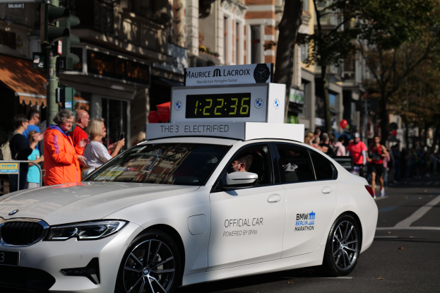 Ein weißer BMW mit einer Uhr auf dem Dach fährt eine Straße entlang, mit einer Person im Auto und einer Gruppe von Menschen am Straßenrand, während im Hintergrund Gebäude, Bäume und Verkehrszeichen zu sehen sind.