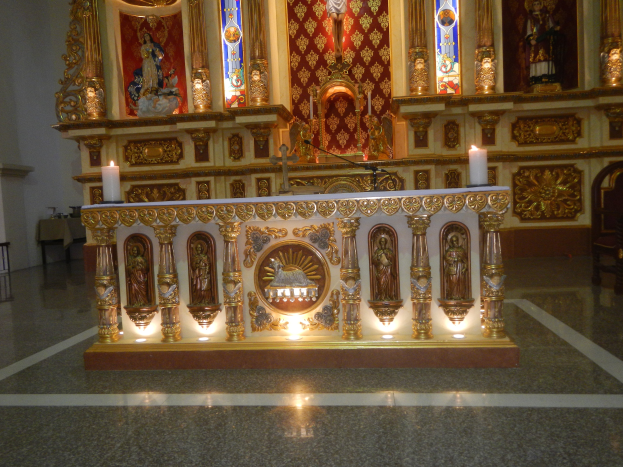 Ein Altar in einer Kirche, beleuchtet von Kerzen, umgeben von Skulpturen, Kerzenständern, einem Mikrofon mit Ständer, einem Stuhl, einem Tisch mit verschiedenen Gegenständen und Buntglasfenstern im Hintergrund.