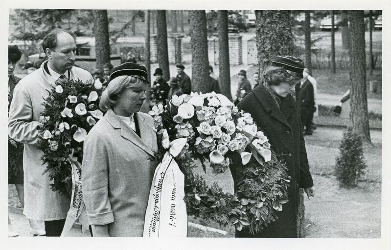 Eine Gruppe von Menschen hält Blumensträuße in den Händen und steht zusammen bei einer Gedenkfeier, mit Bäumen und Gebäuden im Hintergrund, dargestellt in Schwarz-Weiß.