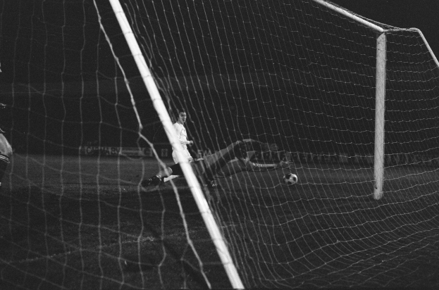 Schwarzes und weißes Foto eines Fußballtorwarts, der einen Ball in der Nähe des Torpfostens abwehrt, mit dem Netz im Hintergrund.