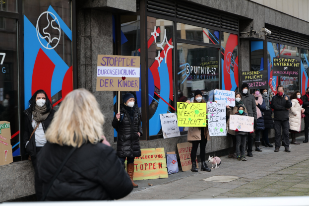 Gruppe von Menschen mit Masken und Schildern vor einem Gebäude mit Glaswänden und -türen, mit Protesttafeln, Kameras und einem Hund im Bild unten.